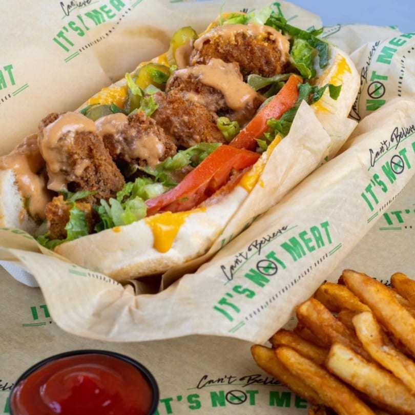 A vegan po'boy sandwich with crispy plant-based protein, lettuce, tomatoes, pickles, and sauce, served with fries and ketchup.