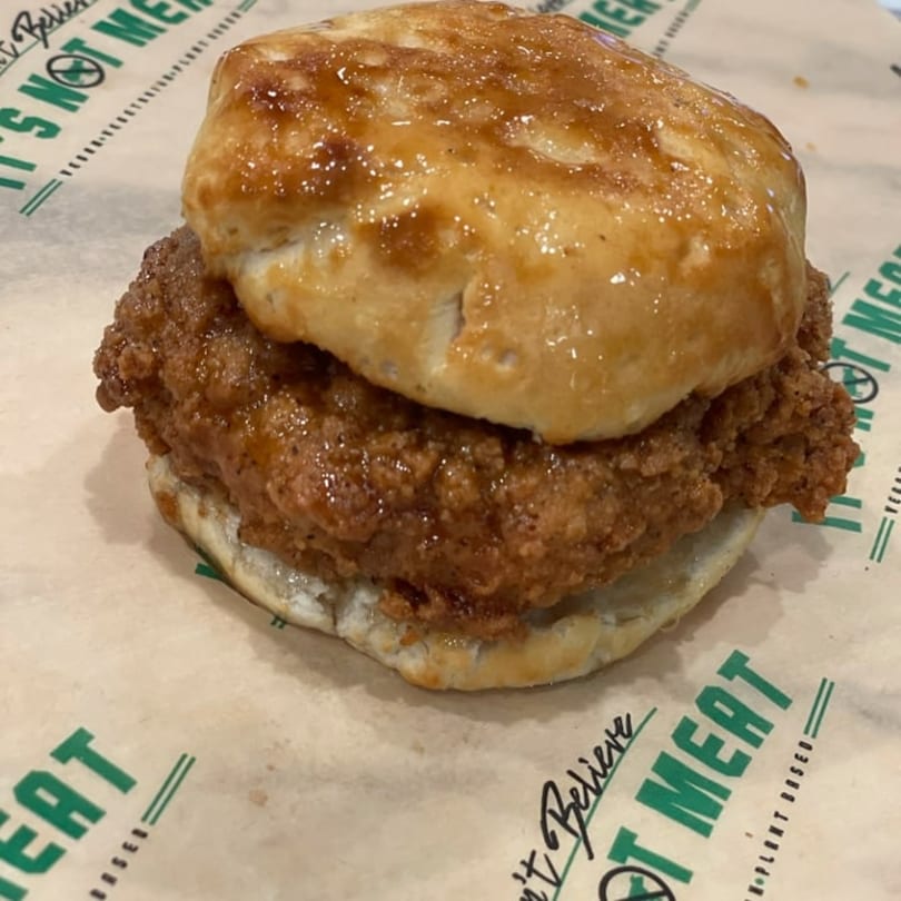 Vegan fried chicken biscuit with a shiny, sweet glaze on branded "Can't Believe It's Not Meat" paper.