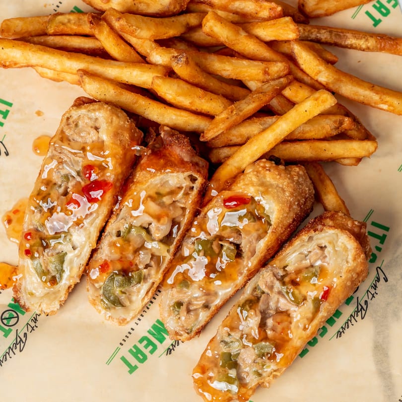 Vegan egg rolls with sweet chili sauce and a side of golden french fries on branded paper.