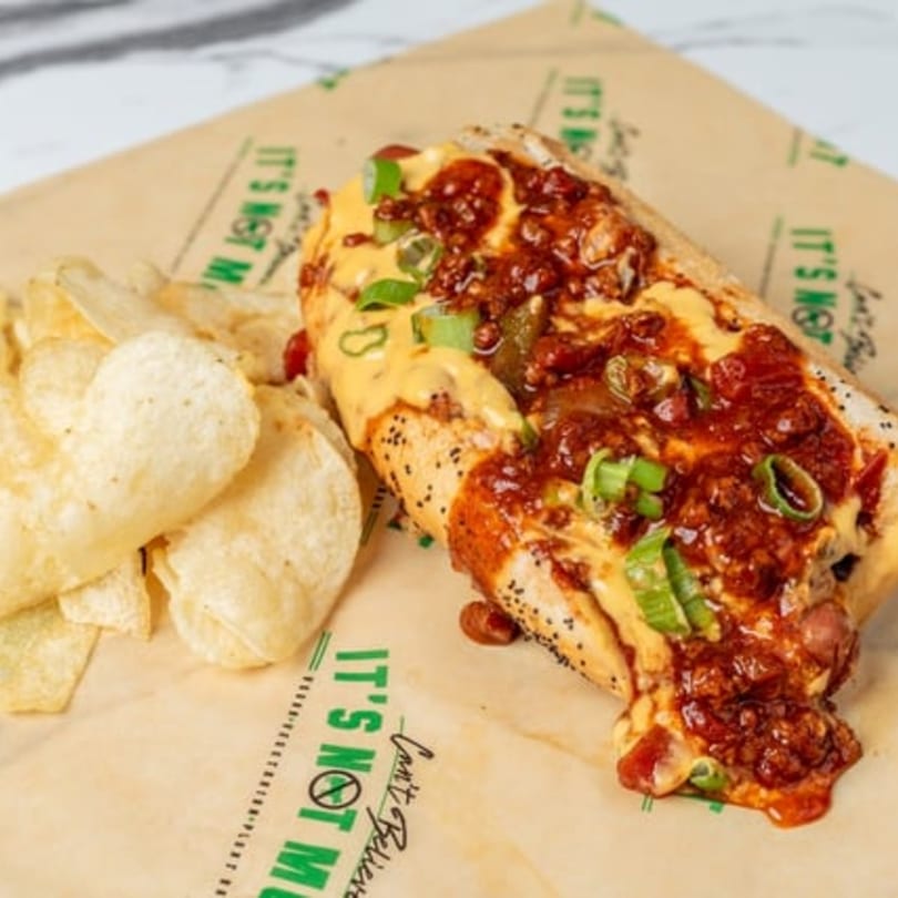 Vegan chili cheese dog with green onions and a side of potato chips on branded paper.
