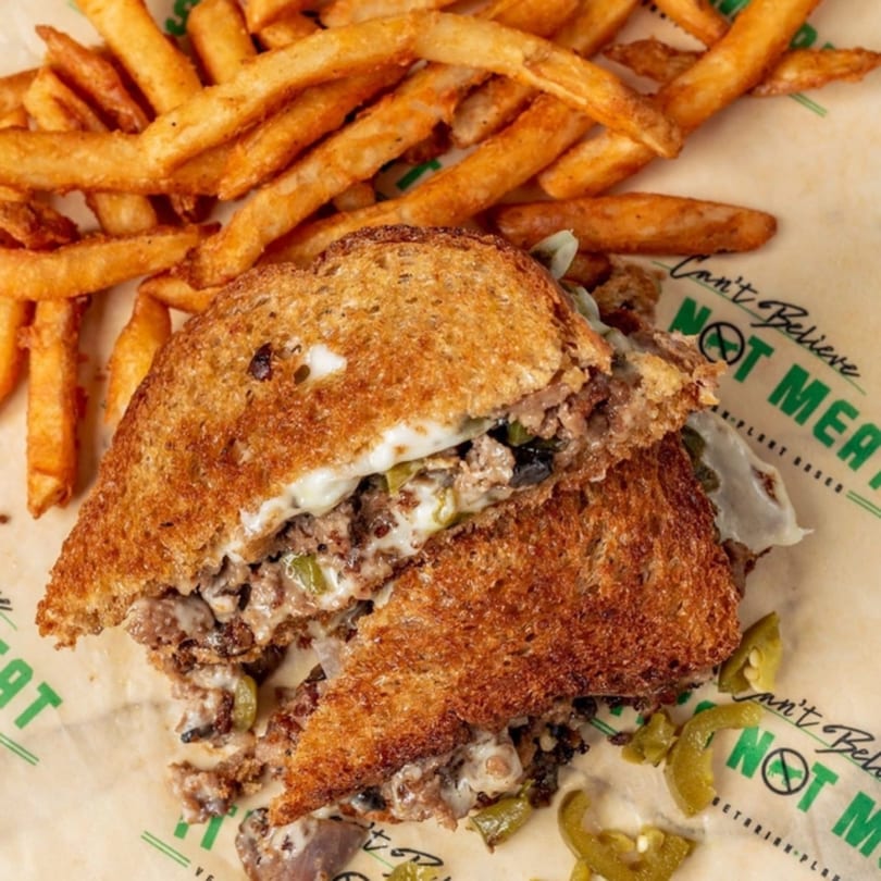 Vegan Philly cheesesteak sandwich, cut in half, with fries and pickled peppers on branded paper.